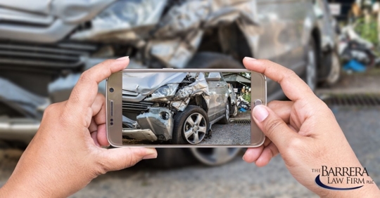 Woman's hands holding a cell phone while taking a picture of a damaged car.