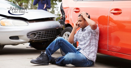 Man sitting on pavement against his car that has been hit by another car.