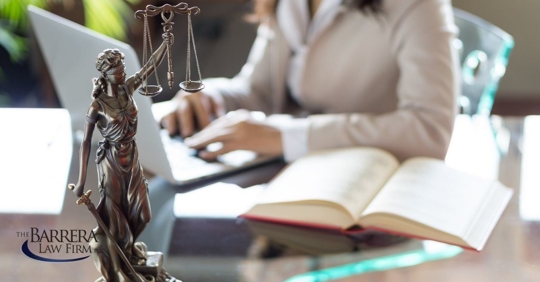 Woman sitting at desk typing on her laptop with a book beside her and a small statue of Lady Justice holding the scales of justice.